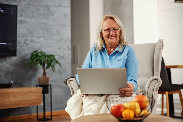 Smiling senior woman sitting at home in her chair with laptop in her lap and hanging on internet.