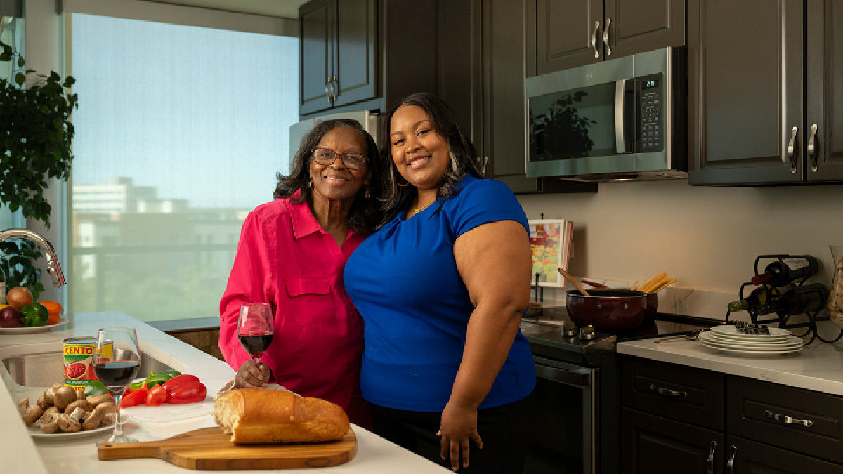 A senior woman and her adult daughter are in the kitchen drinking red wine and cooking