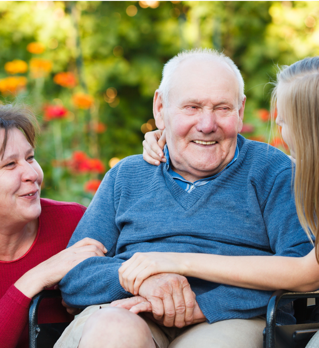 Granddaughter and wife crouch next to an elderly man in a wheel chair to hug him