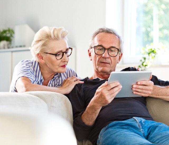 Elderly man sitting on sofa in the living room at home and showing something on digital tablet his wife. Senior woman peeking on screen.