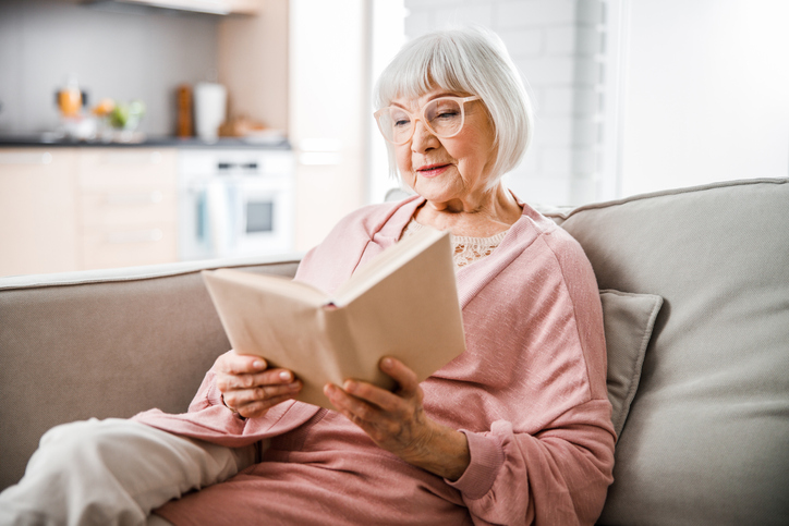 A senior woman sits on a couch reading a book